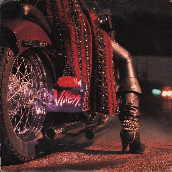 A leather-clad leg wearing a chained high-heeled boot stands beside a motorcycle displaying a Vixen license plate
